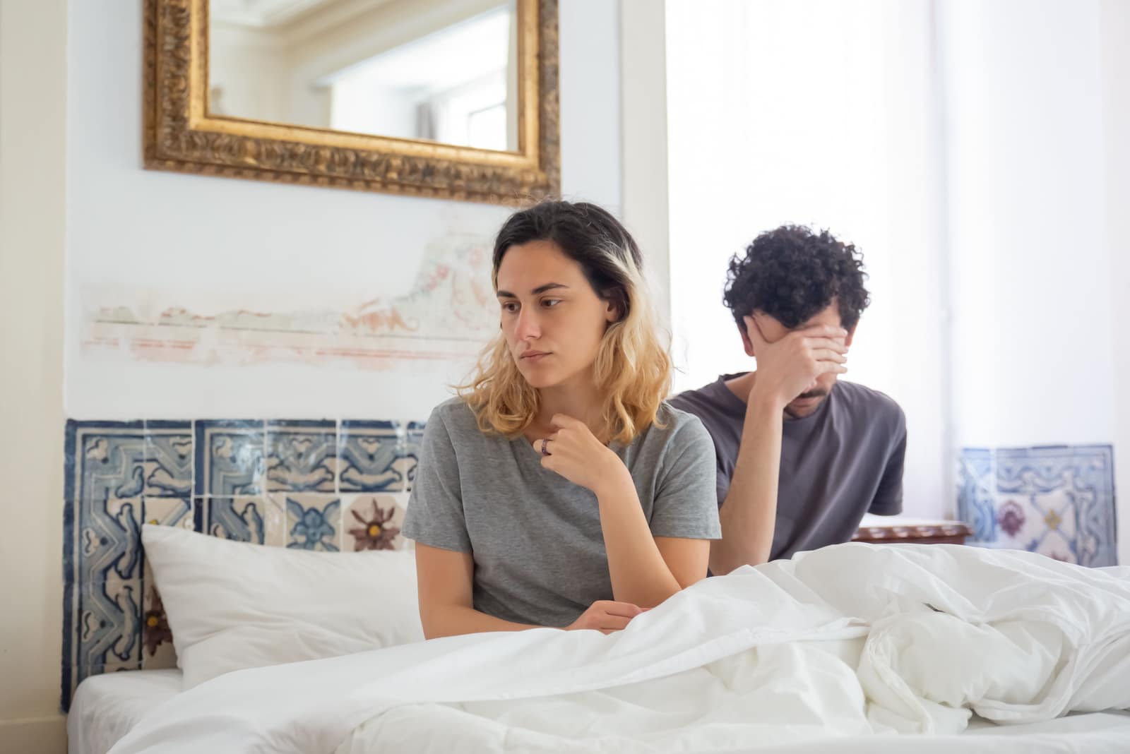 Woman sitting thoughtfully on a bed beside a distressed partner in a bright bedroom